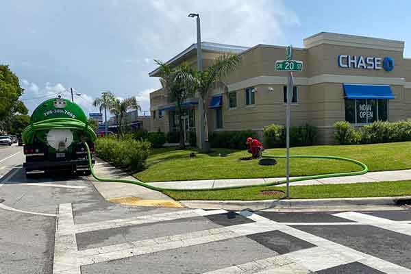 A green septic truck is parked near a Chase bank, with a worker tending to the lawn